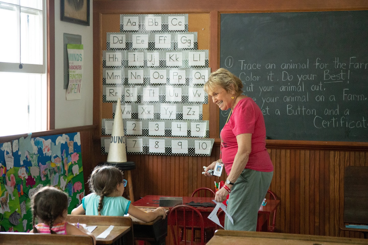Maestra sonriendo mientras ayuda a dos niñas en pupitres en un aula.