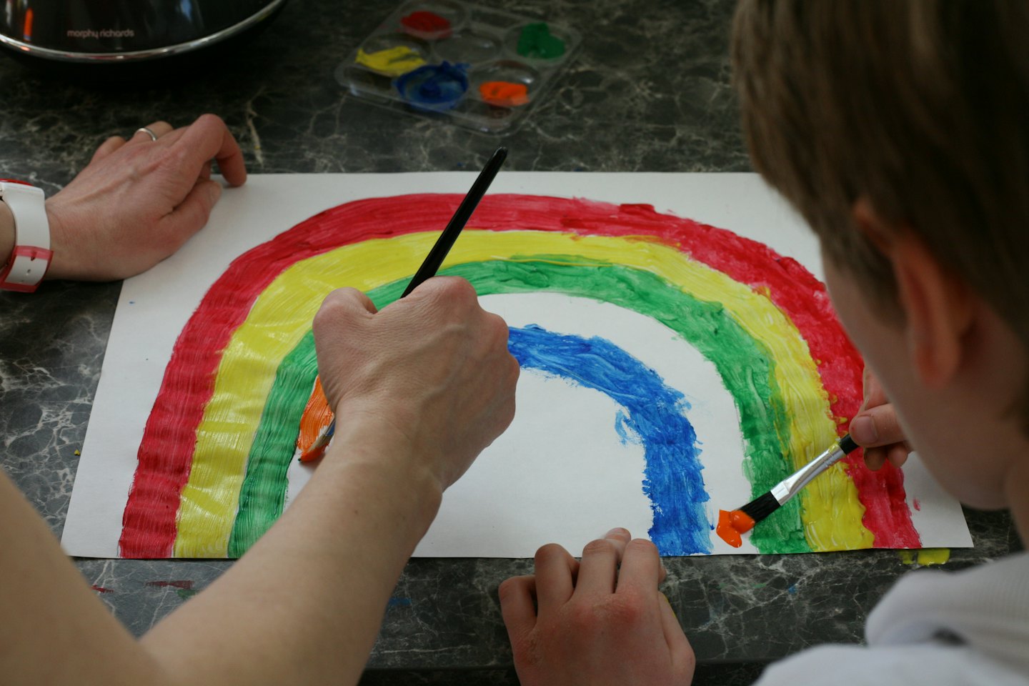 Manos de adulto y niño pintan un arcoíris en papel blanco.